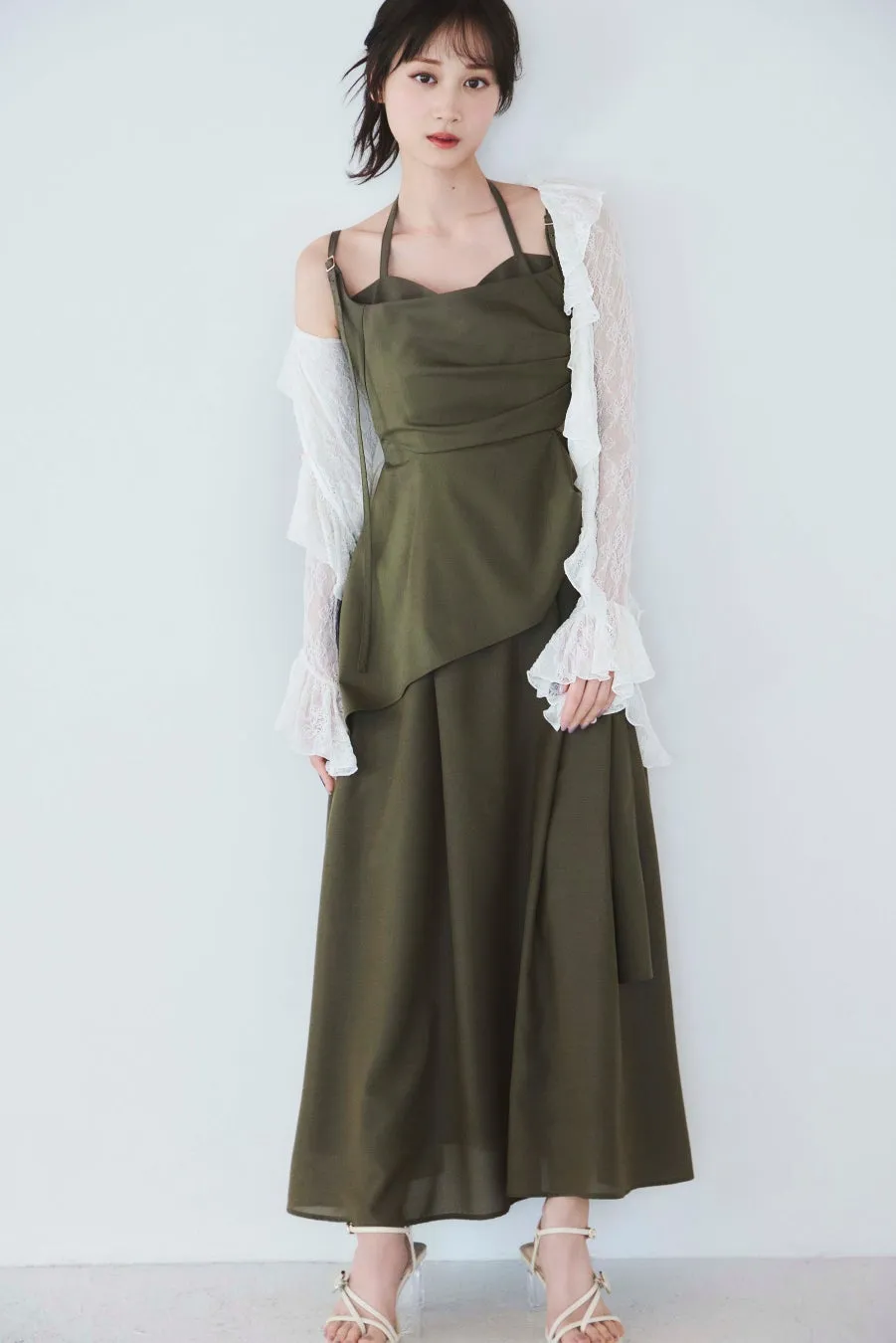 女性が白い壁の前に立っており、オリーブグリーンのドレスと白いレースのカーディガンを着ています。