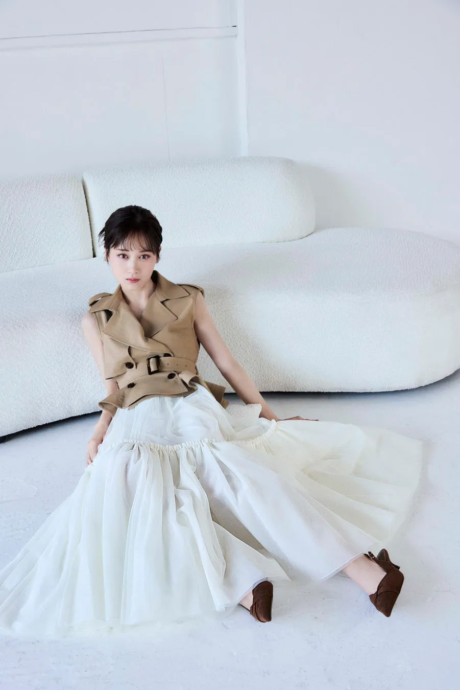 白いソファの前に座っている女性。彼女はベージュのノースリーブのジャケットと白いスカートを着ています。