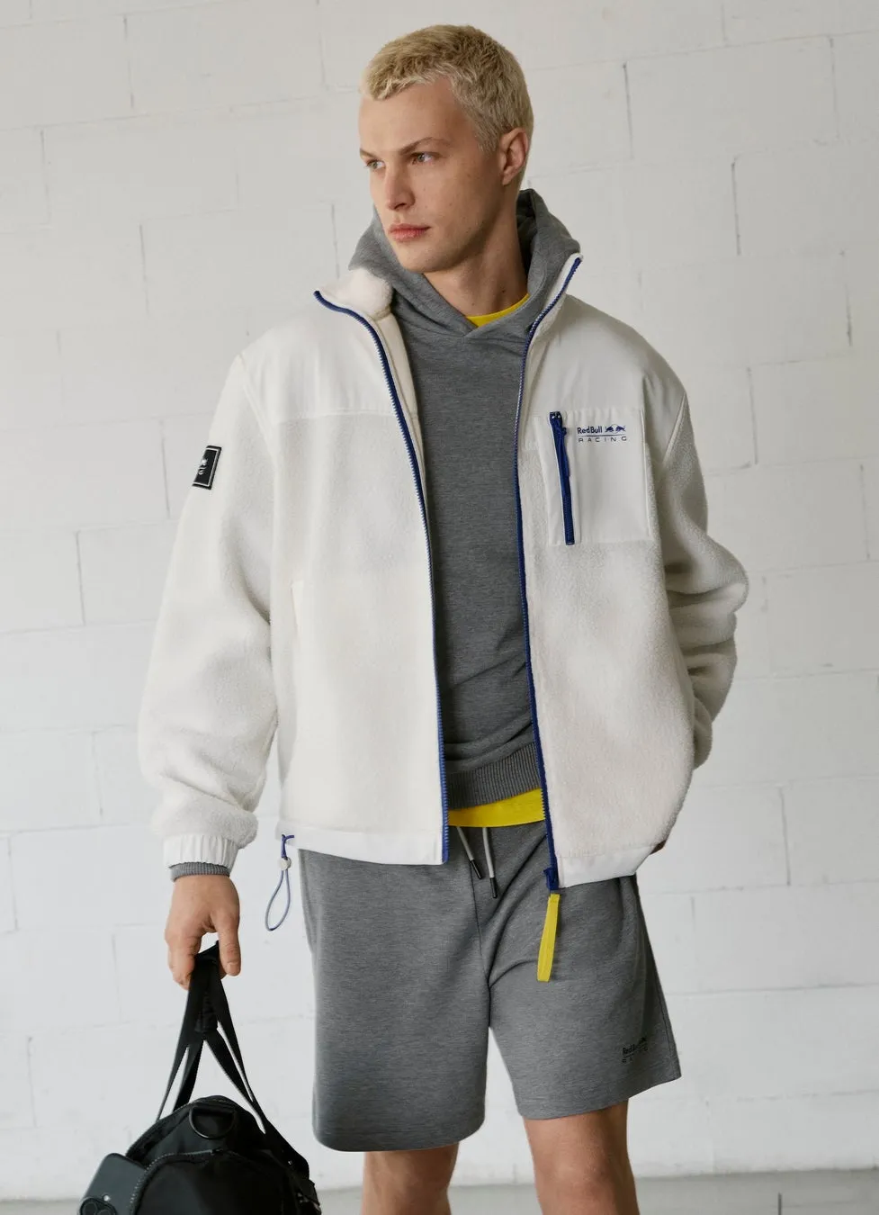 白のフリースジャケットとグレーのパーカーを着た男性。彼は黒いバッグを持っており、グレーのショーツを履いています。