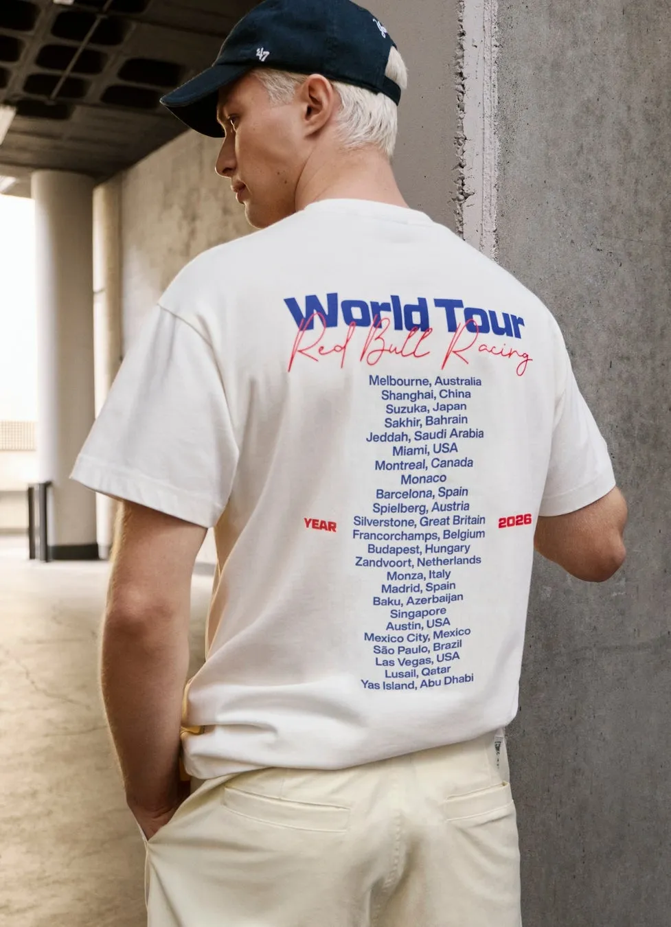 白地のTシャツを着た男性の画像。Tシャツの背面には「World Tour Red Bull Racing」の文字と、世界中のレース開催地がリストアップされています。男性は紺色のキャップを被り、明るい色のズボンを履いています。