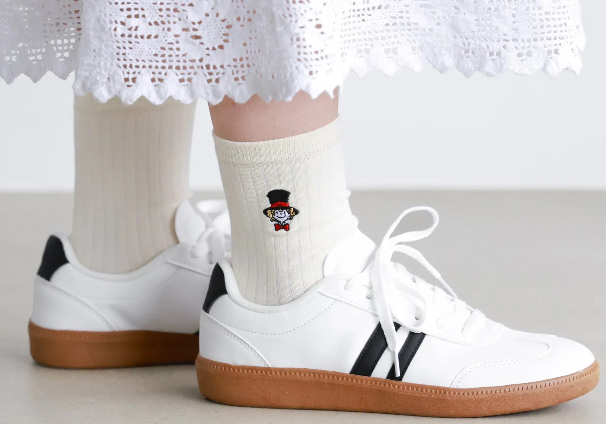 白いスニーカーを履いた足元の写真。白い靴下には、帽子をかぶったキャラクターの刺繍が施されている。背景には白い壁と床が写っている。