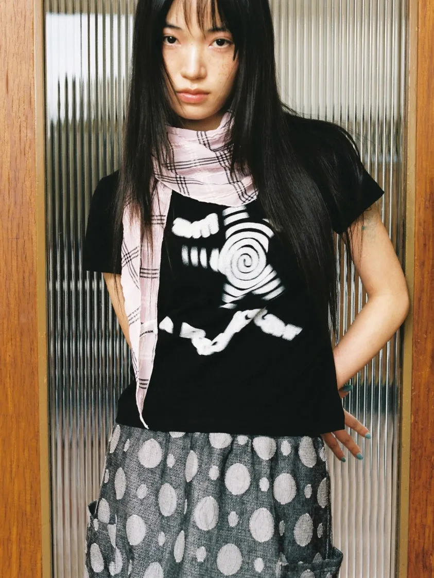 黒いTシャツに模様入りのスカートを着た女性が立っている写真。女性はピンク色のスカーフを首に巻いている。