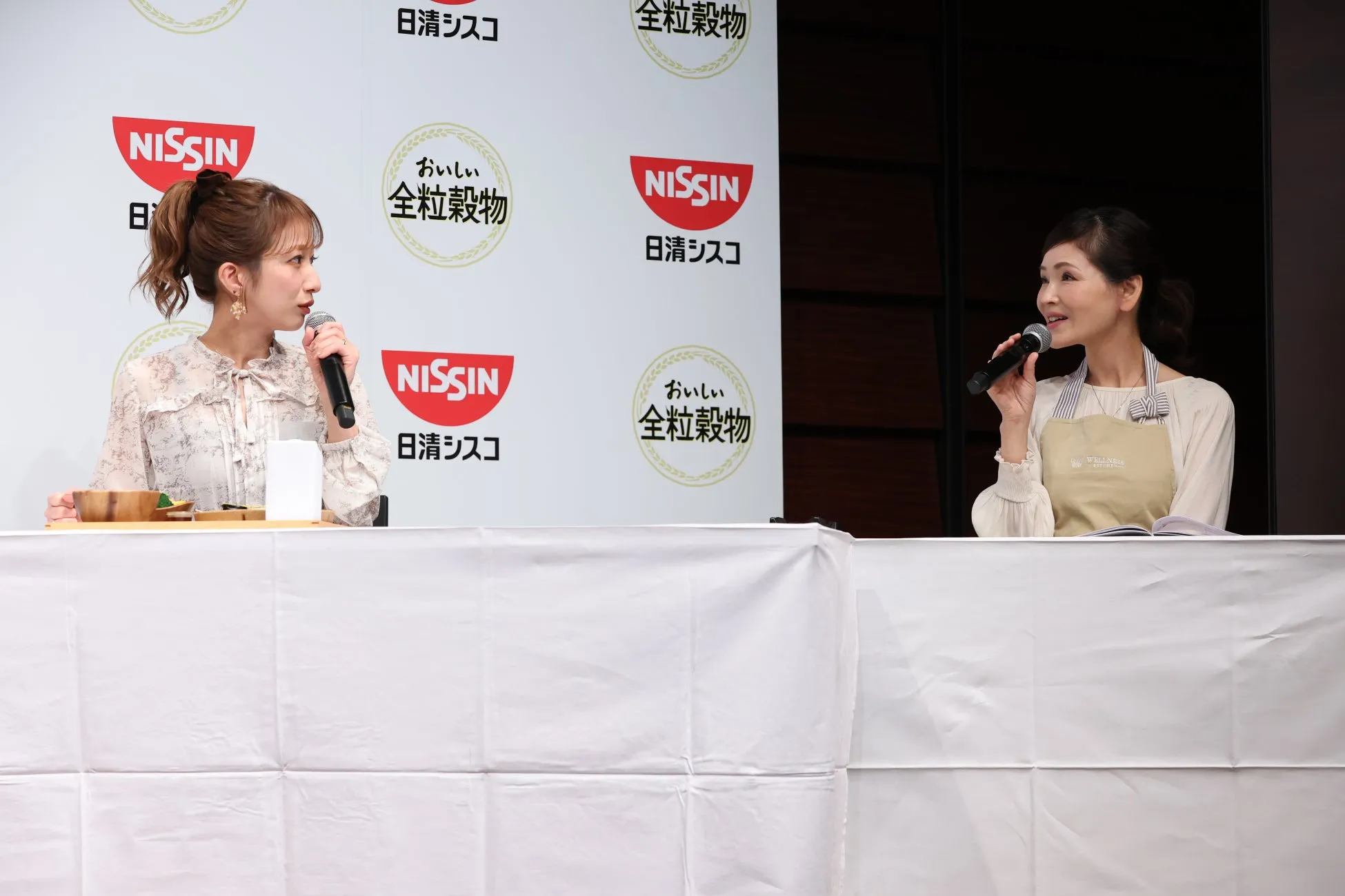 2人の女性がマイクを持って話している様子。背景にはNISSINと全粒穀物のロゴが見える。