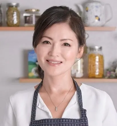 エプロン姿の女性のポートレート。彼女はカメラに向かって微笑んでおり、背景には棚に置かれた瓶やティーポットが見える。