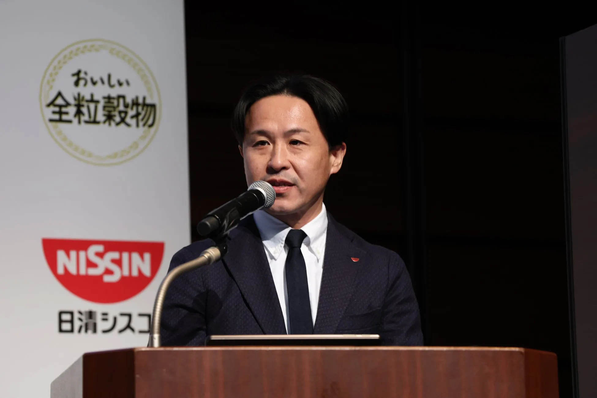 男性がマイクを使って話している様子。背景には「おいしい全粒穀物」のロゴと「NISSIN」のロゴが見える。