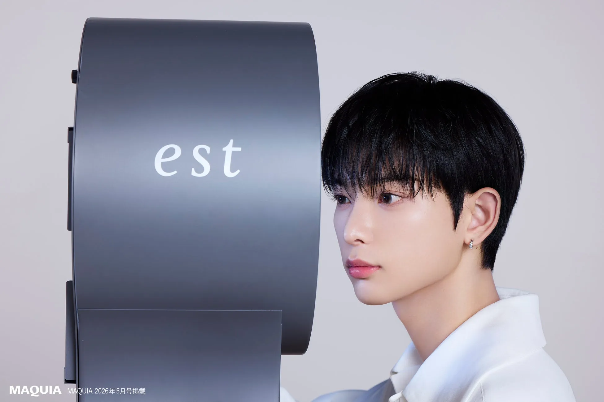 若い男性が、estと書かれた灰色のオブジェクトのそばに立って顔を横に向けているポートレート。男性は白いシャツを着ており、小さなイヤリングをしている。背景は白い。