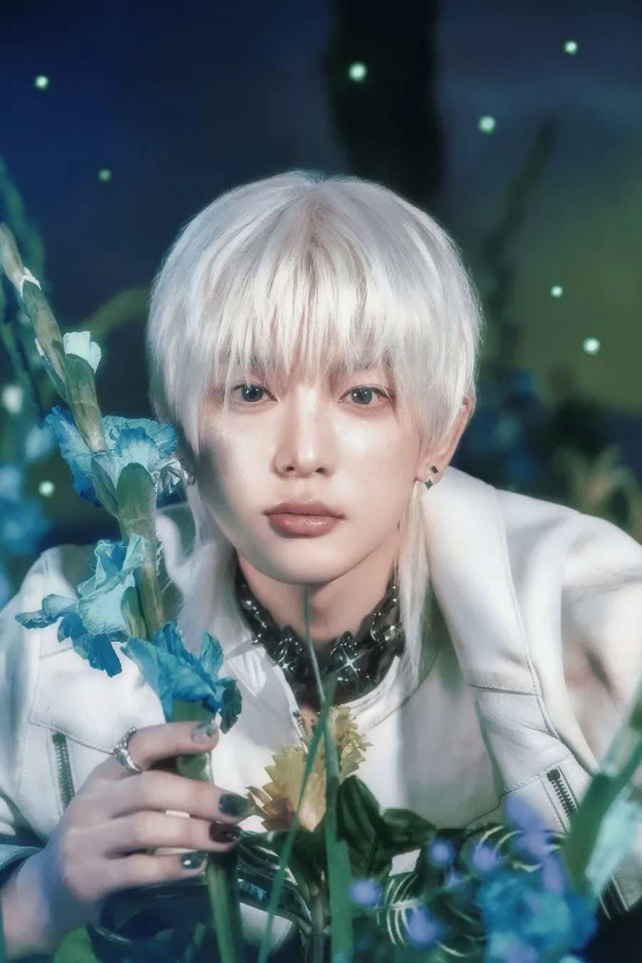 白髪の男性が花に囲まれてポーズを取っている様子を描いた写真。彼は白いジャケットを着ており、顔の近くには青い花が咲いています。背景は暗く、緑の光が散りばめられています。