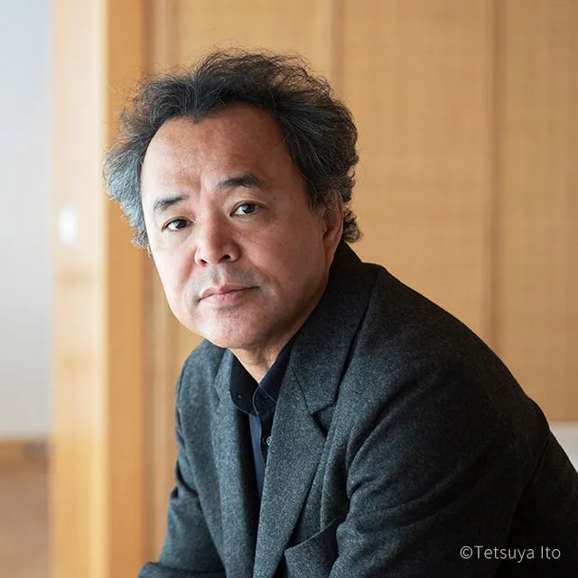 正面を見つめる男性のポートレートです。男性は、グレーのジャケットと濃い色のシャツを着ています。背景は木製の壁です。