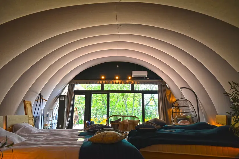 アーチ型の天井と大きな窓が特徴的な、洗練された寝室のインテリア。ベッド、吊り下げ式の椅子、窓の外の緑豊かな風景が、居心地の良い空間を作り出している。