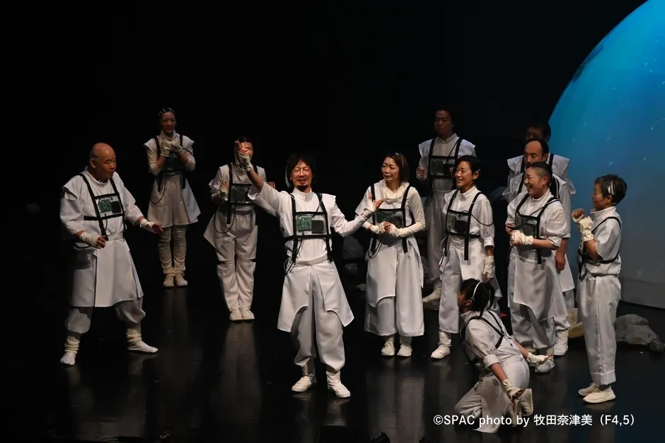 舞台上で白い衣装を着た人々がポーズを取っている写真。中央の男性は両手を広げ、他の人々は彼を取り囲んでいます。背景には青い照明があり、舞台劇のシーンを捉えているようです。