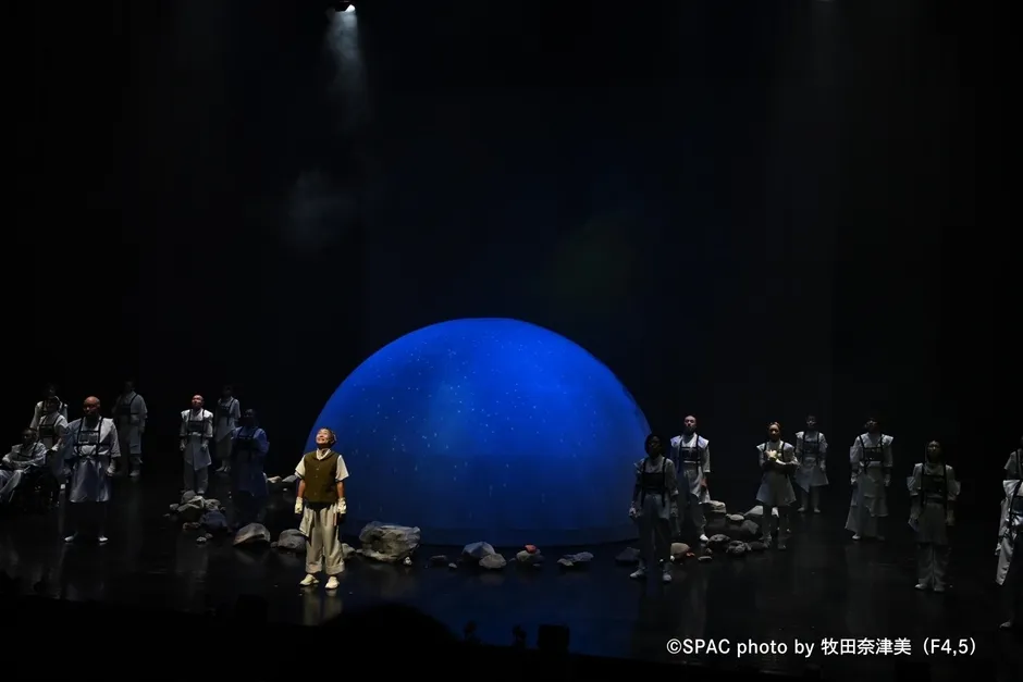 舞台の様子。青い球体のような背景の前で、白い衣装を着た俳優たちが立っている。手前には小石が置かれている。スポットライトが当たっている。