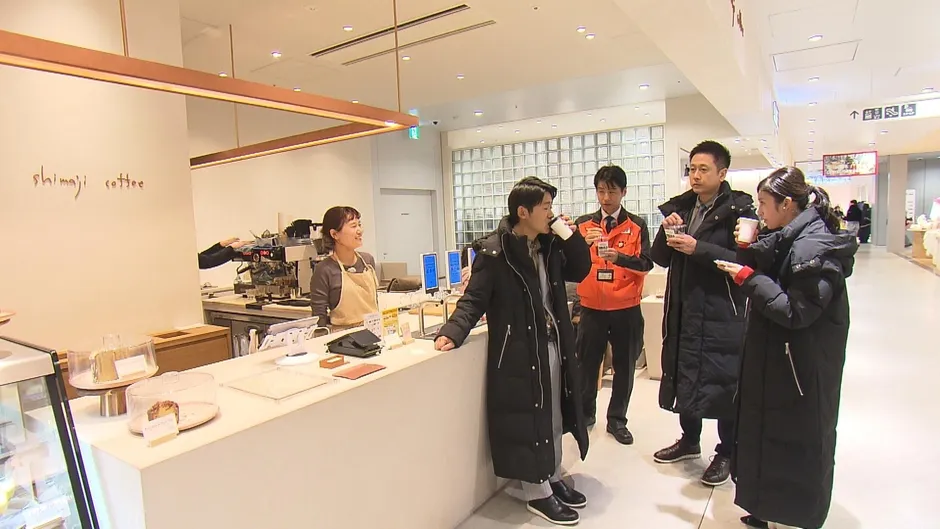 カフェのカウンターでコーヒーを飲む人々の様子を写した写真です。店員が対応し、客はコーヒーを手に会話をしています。