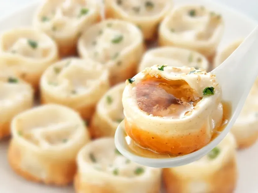 白磁の皿に並べられた点心と、スプーンですくわれた点心のクローズアップ。点心は、丸く、茶色と白の層になっており、上に緑の薬味が乗せられている。点心の中には具材とスープが入っており、スプーンですくわれた点心からスープがこぼれ出ている。