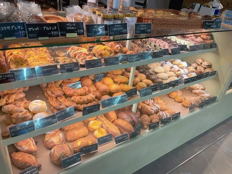 様々な種類のパンが陳列されたショーケースの写真。パンの種類は、クロワッサン、バゲット、デニッシュなど多岐にわたる。価格表示の札も添えられている。