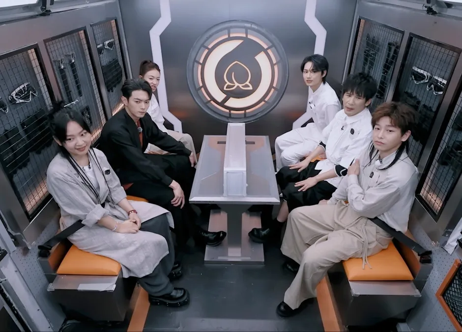 番組セット内で、複数の人々がオレンジ色の座席に座ってカメラを見ている様子。