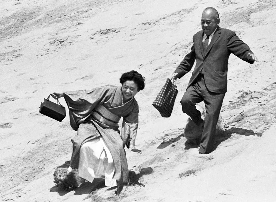 砂の上を歩く男女の白黒写真。女性は着物を着ており、男性はスーツを着ています。二人とも手にバッグを持っています。