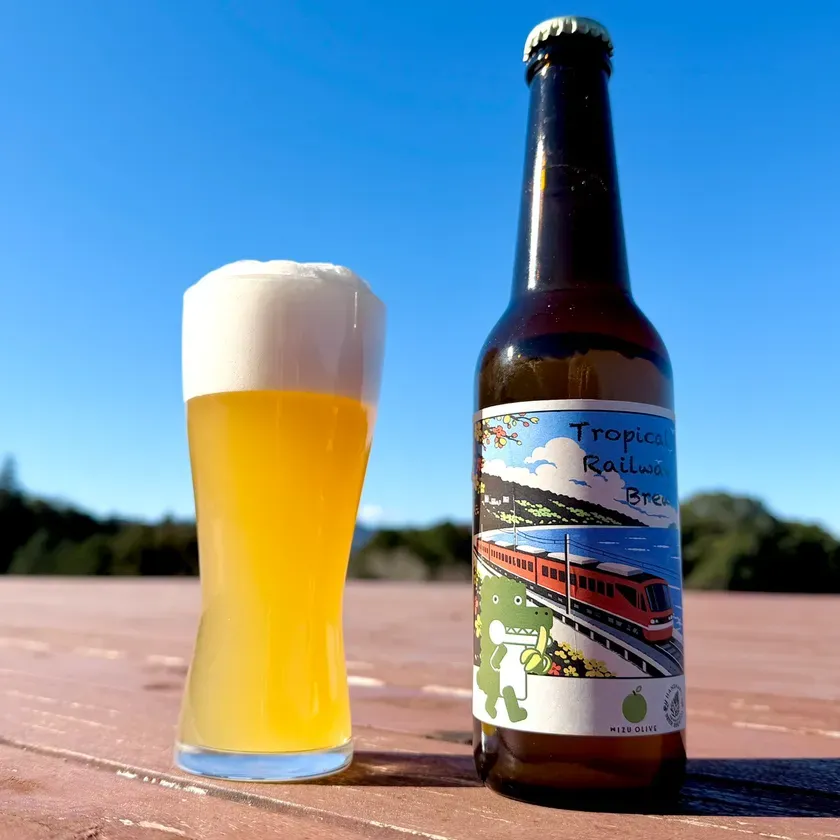 ビールが入ったグラスとビールの瓶が写っている写真です。青空の下、テーブルの上に置かれています。ビールの瓶には、トロピカルな雰囲気のイラストが描かれています。