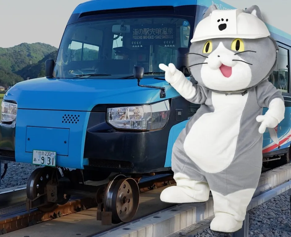 猫の着ぐるみが線路を走るバスの前に立っている様子