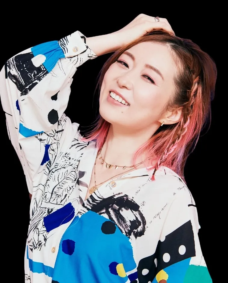 ピンクの髪の女性が黒い背景の前に立って笑顔でポーズをとっています。彼女は白いシャツを着ており、髪は三つ編みで、手は頭に触れています。